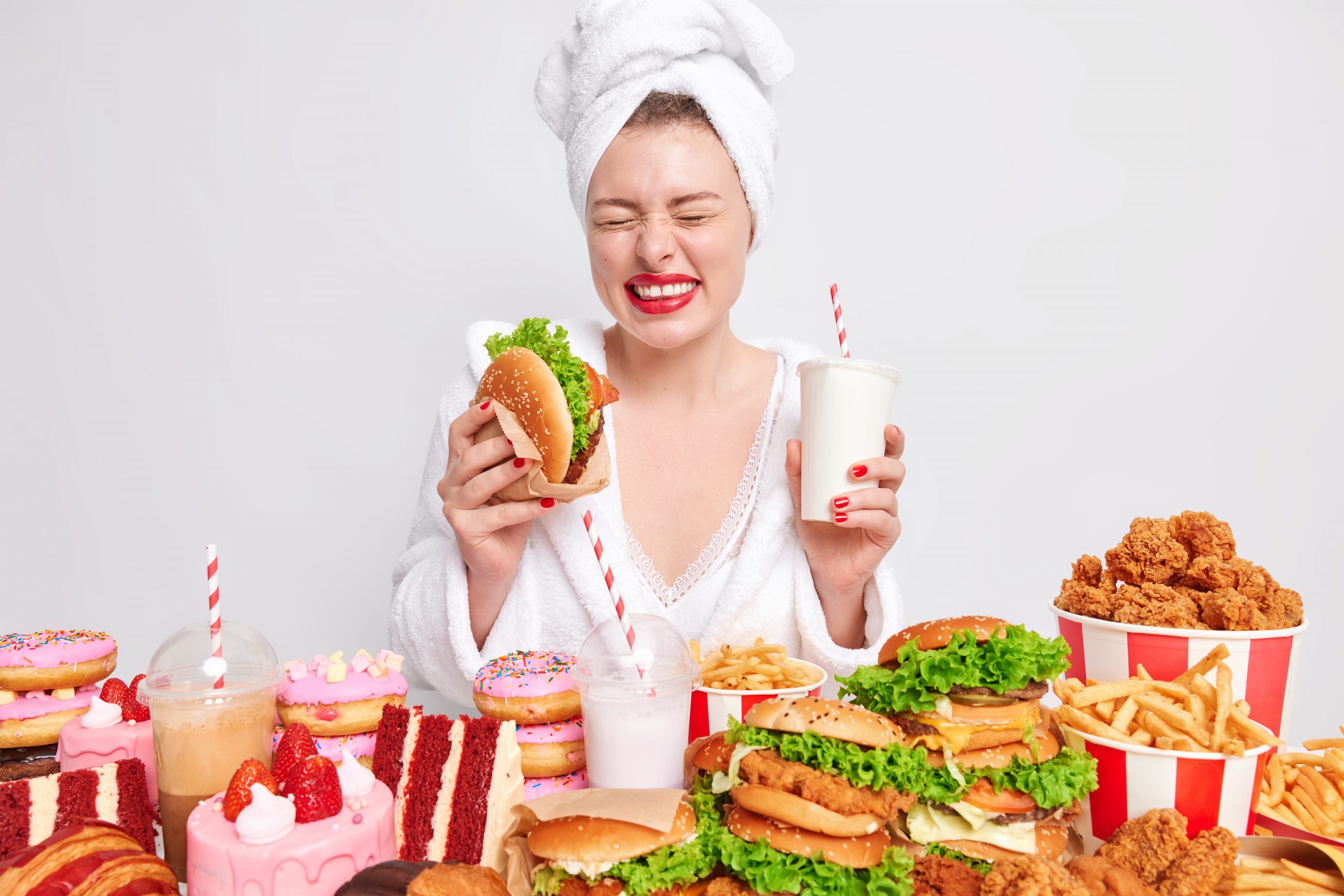 Diet failure and unhealthy lifestyle concept. Overjoyed young woman holds hamburger and fizzy drink wears soft white robe wrapped towel on head prefers junk meal enjoys lunch time. Much calories