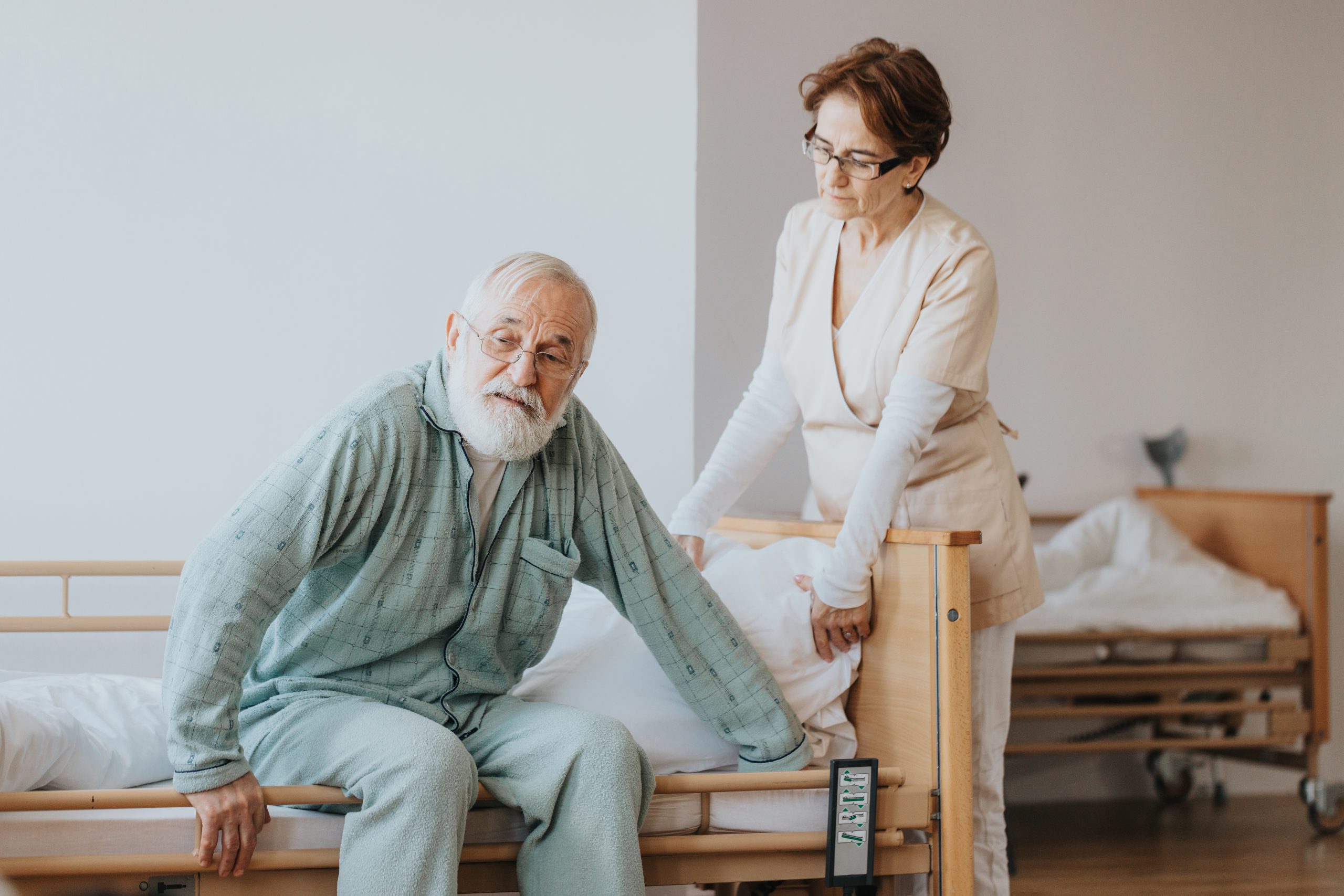 helpful-nurse-beige-uniform-helps-patient-blue-pajamas-getting-up-from-bed-scaled.jpg